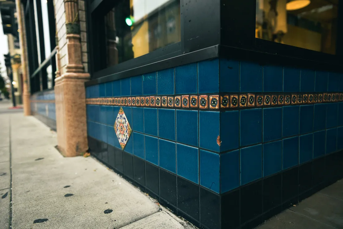 The tiled corner of a Napa building, deep blue glazed tile with a painted ceramic diamond and a band of patterned border tiles above it, meeting the pavement.