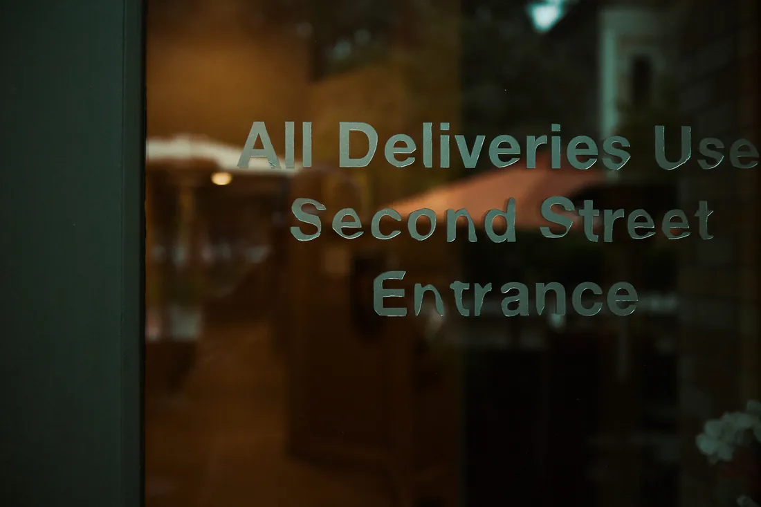 Lettering on a glass door reads: all deliveries use Second Street entrance, with a warmly lit room out of focus behind the glass.